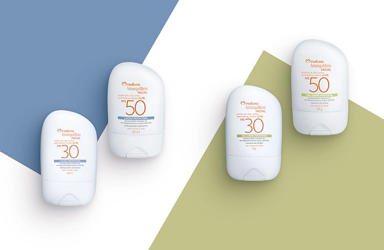 Foto de cuatro protectores solares Fotoequilibrio de Natura sobre fondo geométrico azul, blanco y verde. Dos envases con FPS 30 y dos con FPS 50, todos en formato de frasco blanco con detalles en naranja y azul o verde.